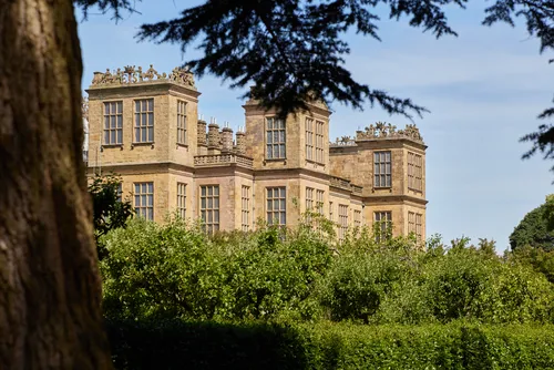 This architectural photograph depicts Hardwick Hall, a prominent Elizabethan manor house located in Derbyshire, United Kingdom. Taken during the morning in the summer season, the image shows Hardwick Hall’s distinctive façade partially obscured by surrounding trees and greenery, with clear blue skies overhead. The manor house is celebrated for its grand architecture and extensive windows, and is managed by the National Trust. As a landmark of English heritage, Hardwick Hall exemplifies Elizabethan design and is situated within the scenic countryside of Derbyshire.
